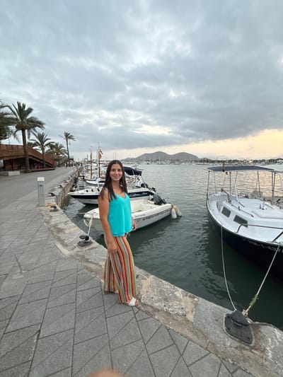 A full-body, eye-level, outdoor shot features a young woman posing on a paved promenade in Port D'alcúdia, Spain. The woman, with long, dark hair and a bright turquoise sleeveless top, stands on a walkway made of light gray interlocking pavers. She is wearing colorful, wide-leg striped pants in red, orange, yellow, and green hues, and white platform sandals. Her hands are at her sides, and she is looking directly at the camera with a smile.
Behind her, the promenade transitions to a stone-lined harbor where several small white boats are docked. The water is dark green. In the background, a larger white boat with a dark canopy is prominent. Beyond the boats, a dense collection of sailboats with white masts fills the horizon, extending towards distant, hazy mountains. The sky is overcast with soft gray clouds, and a sliver of pale orange is visible on the far right, suggesting either dawn or dusk. Palm trees and a wooden structure with a curved roof are visible on the left side of the frame, adding to the Mediterranean atmosphere. The overall lighting is dim and diffused.