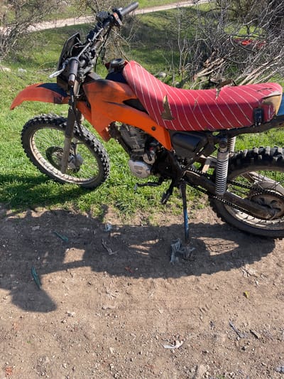 An orange dirt bike with a heavily worn, red and white striped seat is parked outdoors in Topchii, Bulgaria, on a sunny day. The motorcycle features knobby tires, an exposed engine with "HONDA" visible on its casing, and a blue and black kickstand supporting it. The front fender and tank shrouds are orange, and the front wheel has a disc brake. The bike rests on a patch of uneven dirt and small gravel, with scattered pieces of white plastic debris, a green rope, and other detritus on the ground. Beyond the dirt, an area of vibrant green grass is visible, followed by numerous bare, thorny bushes or small trees. In the far background, an unpaved road or path traverses a green landscape. No individuals are present in the scene.