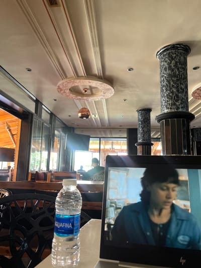 An indoor scene shows a laptop displaying a person on screen, positioned in the foreground. To the left of the laptop, an Aquafina water bottle stands on a white surface. Behind the laptop, a black patterned chair is visible. The background reveals a dining area with wooden tables and chairs, and large windows looking out to a green outdoor space. The ceiling features decorative elements, including columns with a marbled pattern and circular light fixtures. The location is identified as El Hajeb, Morocco.