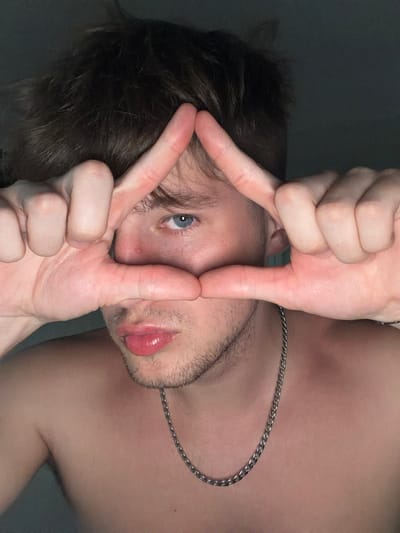 A close-up, low-angle, indoor portrait shows a young person with light brown hair and blue eyes, framing their face with their hands. Their right hand is placed over their forehead, with the index and middle fingers extended upwards, and their left hand is placed below their nose, with the index finger extended horizontally. The person is looking directly at the camera. They are shirtless, wearing a silver chain necklace. The background is dark and nondescript. The scene is consistent with a self-portrait taken in a residential setting. No specific location in Vanino, Russia, can be determined from the image.