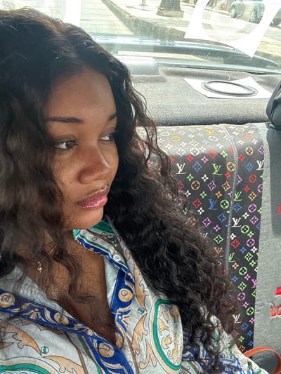A woman is lying down in a vehicle. Her head rests on a black Louis Vuitton pillow with multicolored logos. She is wearing a patterned shirt with blue, green, and white colors. Part of the vehicle's window is visible, showing a blurry cityscape of Rumuigbo, Nigeria, suggesting the scene is inside a moving or parked car in the area.