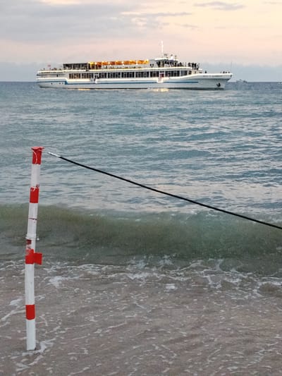 A wide shot captures a calm body of water with a large, white passenger ship illuminated by interior lights sailing in the distance under a muted, cloudy sky. In the foreground, a red and white striped pole stands planted in the shallow water near a sandy shore, with a dark rope extending diagonally across the frame from the pole towards the right. The water is gently rippled, with small waves lapping at the shore. No specific location in Bosteri, Kyrgyzstan, can be identified from this image.