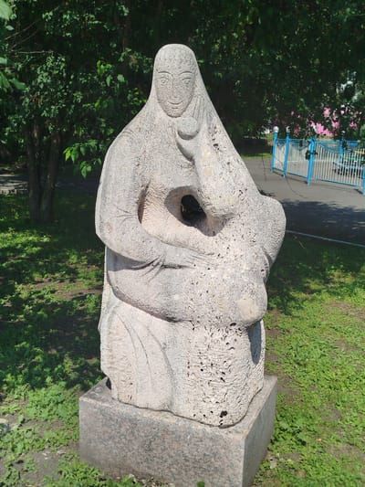 A light-colored stone sculpture of a seated woman holding a baby is positioned outdoors on a granite pedestal in a grassy area. The sculpture depicts a figure with a headscarf, an abstract face with carved features, and draped clothing. The woman's arms are wrapped around a smaller, rounded form representing a child. The sculpture has a rough, textured surface with visible pores and imperfections. Behind the sculpture, a paved walkway and a blue and white metal fence with pink structures beyond are visible. Lush green trees and foliage surround the immediate area, with dappled sunlight casting shadows on the ground. This sculpture is located in the city of Yalutorovsk, Russia.