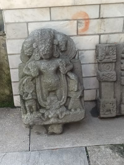 The image shows a stone sculpture of a figure with multiple heads, possibly a deity, situated outdoors. The sculpture is partially weathered and exhibits intricate carving. To the right of the main sculpture, another stone carving is visible, appearing to be part of a pillar or panel with decorative motifs. The background consists of a brick wall made of rectangular blocks, some of which are lighter in color than others. A faint orange circular mark is present on one of the lighter bricks. The ground is paved with stone slabs, with some green vegetation visible in the lower left corner and between the slabs. The overall setting suggests an archaeological site or a display of historical artifacts. The location is identified as Gojra, India.