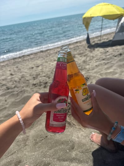 Two people are holding up two bottles of Somersby cider, one raspberry and lime and the other mango and lime, in a toast on a beach in Aheloy, Bulgaria. The beach has dark sand and the Black Sea is visible in the background. A yellow beach umbrella and a tent are also visible behind the people. The people are wearing bracelets. The sun is shining brightly, and the water is calm with small waves.