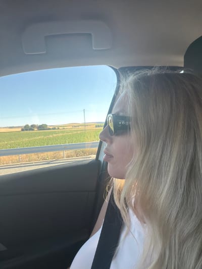 A medium close-up shot shows a woman with blonde hair wearing black-rimmed sunglasses with "VOGUE" printed on the side. She is in the passenger seat of a car, looking out the window. The car's interior is visible, including the door panel and a black seatbelt. Outside the window, a sunlit rural landscape is visible, featuring green fields, a road with a guardrail, and distant trees under a clear blue sky. The scene appears to be in Villarrubio, Spain, suggested by the landscape and lighting.