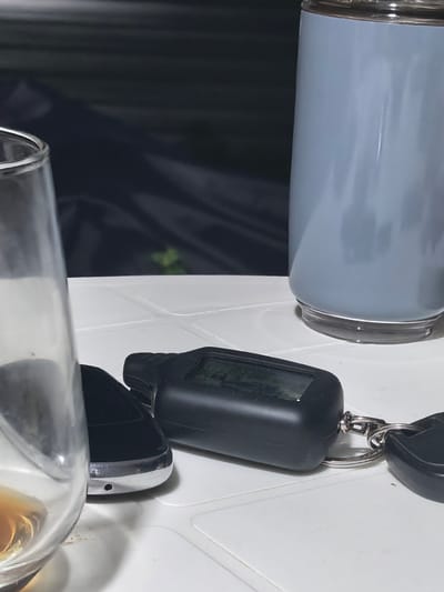 An outdoor table is laden with objects, suggesting a casual setting. In the foreground, a clear glass partially filled with a brownish liquid, possibly alcohol, is situated on the left. To its right, a black car remote key fob with a small screen and buttons lies on the white surface, attached to a keychain with a silver ring and another black object. Behind these, a tall, light blue tumbler stands upright. The background is dark and out of focus, hinting at foliage and possibly a dark tarp or fabric. The overall scene is still and appears to be captured in low light conditions. The location is identified as Beloyarskaya Zastava, Russia, though no specific urban landmarks are visible.
