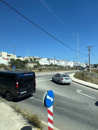 This is a medium shot, outdoors, sunny day, eye-level perspective, of a road in Thira Municipal Unit, Greece. In the foreground on the left, a black van is parked, facing right, with its rear towards the viewer. The van has a license plate that reads "EMT-3874". The side of the van has text that reads "TRAVEL MAN E.E.". To the right of the van, a silver sedan is driving away from the viewer, also facing right, and has a license plate that reads "ZK8-4518". In the middle of the road, there is a blue and white circular directional sign with a white arrow pointing right. Further down the road, there is a white sign with the word "Camping" on it. In the background, there are white buildings with blue accents, characteristic of Greek architecture, on a hill. There are also telephone poles and power lines throughout the scene. The sky is clear and blue.
