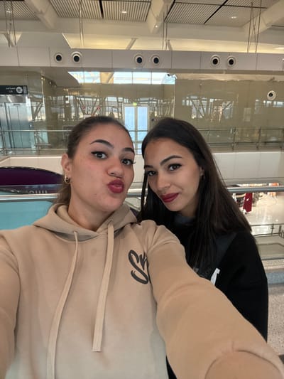 Two young women are captured in a selfie, likely inside a modern airport terminal. The image is taken from a low angle, showing their faces close to the camera against a bright, contemporary architectural background.

The woman in the foreground, to the right, wears a light beige or tan hoodie with a black, stylized logo on the chest. She has a playful expression, pouting her lips towards the camera, and her face is adorned with freckles and dark red lipstick. A small gold hoop earring is visible in her right ear.

Behind her, to the left, another woman with long dark hair and a black top or hoodie smiles subtly towards the camera. She also wears dark red lipstick and has well-defined eyebrows.

The background features bright, clean architecture with large glass panels, metal beams, and white walls and ceilings. Circular light fixtures are visible along vertical pillars. Reflections in the glass show parts of the interior and what appears to be the blue sky and possibly trees through distant windows, indicating it is daytime. On a column in the background, a sign with white text against a dark background is visible, displaying both Arabic script and the English word "LIFT".

The overall scene suggests a casual and friendly moment, possibly while traveling.