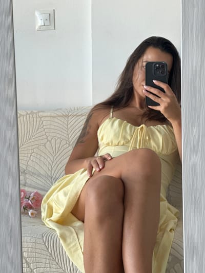 A woman is sitting on a couch, taking a selfie in a mirror. She is wearing a yellow dress and has long, dark hair. There are some flowers on the couch next to her. The wall behind her has a light switch.