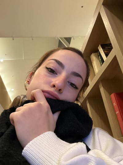A young woman poses, head tilted, with a contemplative expression. Her hand is near her mouth. She is wearing a white sweater and a black scarf. The background includes a wooden shelf with books and a bright white wall with mirrored panels. The lighting is soft and warm. The image is taken from a low angle.