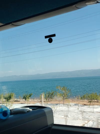 The image captures a scenic view from inside a vehicle, looking out through a window onto a vast natural landscape. The perspective is from a passenger's seat, showing a dark-colored dashboard or seat in the foreground, where a blue water bottle and some white items are placed on the left side.

Beyond the window, a serene body of water, likely a lake or sea, dominates the middle ground with its deep blue-green hues and gentle ripples. On the far side of the water, a range of distant mountains or hills stretches across the horizon under a clear, bright blue sky. Several thin, dark power lines horizontally traverse the upper portion of the sky, adding an element of human infrastructure to the natural scene. A small black rectangular object with a circular attachment is affixed to the window pane, visible against the sky.

Between the vehicle and the water, a strip of land is visible. It features sparse green vegetation, including small trees and bushes. A significant portion of this shoreline appears to be covered with a light-colored material, possibly white plastic sheeting used in agriculture or landscaping.

The photograph depicts a sunny day, likely during late morning or afternoon, given the bright, even illumination. The overall atmosphere is calm and peaceful, suggesting a journey or moment of appreciation for the natural environment. Given the provided context, this tranquil scene is observed from a vehicle in Iznik, Turkey, overlooking Lake Iznik. No discernible text is visible in the image.