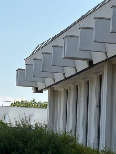 The image captures an unusual upward-facing perspective of an architectural structure, likely a building's overhang or roofline, set against a clear, pale blue sky. The structure features a repeating pattern of white, rectangular panels that are staggered and angled, creating a sense of depth and a visually dynamic facade. These panels appear to be made of a solid material, possibly wood or a composite, and show some signs of wear and tear, with subtle scuff marks and slight discoloration.
Beneath this patterned overhang, a more industrial-looking section of the structure is visible, composed of white, horizontal slats with dark grey or black channels running between them. This part of the building has a functional, possibly ventilation-related, appearance.
To the right of the frame, a dense, vibrant green bush or tree partially obscures another white building. This secondary building has a smooth, uninterrupted surface, punctuated by three small, white rectangular protrusions spaced evenly across its visible area. These protrusions could be vents or decorative architectural elements. The greenery suggests an outdoor setting, possibly a garden or a landscaped area adjacent to the buildings.
The lighting suggests it is daytime, with no strong shadows indicating a specific time of day like sunrise or sunset. The sky is uniformly light, implying fair weather. The overall impression is one of modern or perhaps slightly weathered architecture, with a focus on geometric patterns and contrasting textures. There is no text visible in the image, and no people or specific activity are depicted. The setting appears to be Rungis, France, as indicated by the location context.