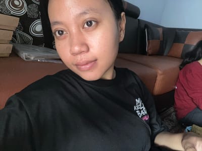 A young woman, appearing to be of Southeast Asian descent, is captured in a close-up, front-facing selfie. She has medium-toned skin, dark hair pulled back, and is wearing a black t-shirt with white and pink text that is partially visible. Her expression is neutral. The background consists of a brown leather sofa with dark grey and brown checkered pillows. To the left of the sofa, there is a stack of brown cardboard boxes and a plastic-wrapped item. The overall setting suggests an indoor environment, possibly a living room. The lighting appears to be from an artificial source, casting a warm glow on the subject.