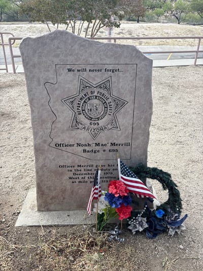In the city of Dragoon, United States, a memorial stone stands in an outdoor setting. The stone is engraved with the outline of Arizona and the Department of Public Safety badge, along with the inscription "We will never forget..." and "Officer Noah 'Mac' Merrill Badge # 695". Below this, text reads, "Officer Merrill gave his l in the line of duty on December 1978, West of this location at mile po 313.6". At the base of the stone, a wreath and artificial flowers are placed. Two small American flags are positioned in front of the wreath and flowers. The ground surrounding the memorial is covered with gravel and dry vegetation, with a metal railing and trees visible in the background.