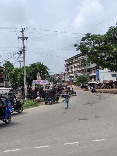The image captures a street scene in Benapole, Bangladesh, under an overcast sky. A paved road occupies the foreground, with a white dashed line marking the lane. Several auto-rickshaws, a common mode of transportation in the region, are present. One blue auto-rickshaw is parked on the left side of the road, while a green one is driving in the right lane. In the middle ground, a man in a blue and white patterned shirt and blue jeans walks away from the camera, carrying two green bags.

Along the sides of the road, a variety of buildings are visible. On the left, there are smaller structures with tin roofs and a prominent utility pole with a dense network of wires. Further down the street, a multi-story building with several floors and numerous windows stands prominently, its facade showing signs of construction with scaffolding. There are also smaller shops and stalls lining the road, displaying various goods.

The background features lush green trees, their foliage dense against the cloudy sky. Several signs are mounted on poles, including one advertising "Medi Inn." The overall impression is that of a busy urban environment with elements of commerce and everyday life. The presence of multiple auto-rickshaws and pedestrians suggests ongoing activity in this part of Benapole.