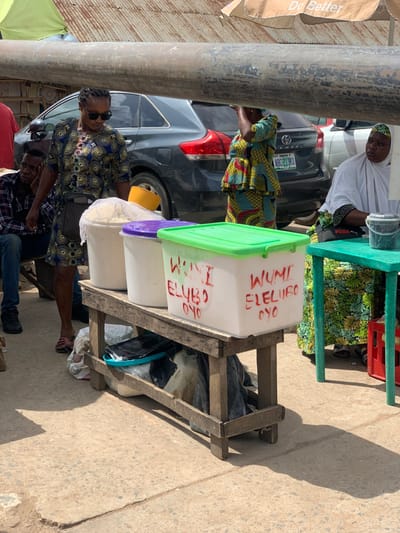 The image captures a street scene in Ondo, Nigeria (latitude: 7.251, longitude: 5.196), featuring a small, open-air market stall. A woman in a blue and black patterned dress stands behind the stall, holding a bag over her shoulder and looking toward the left of the frame. Two large white plastic containers with green and purple lids are on a wooden table in front of her. The containers are labeled "Woni Elubo Oyo" and "Wumi Eleubo Cyo" in red marker. A woman in a floral patterned dress stands behind the stall, partially obscured by a large metal pipe. She is looking towards the camera. A third woman in a white head covering sits to the right of the stall, holding a small metal container. The street is paved with concrete and there is a gray car parked to the right of the stall. The environment is bright and sunny. A man in a white shirt and dark pants sits on the edge of the pavement, looking at the woman behind the stall. The scene is bustling with activity, and the women appear to be selling a variety of goods.