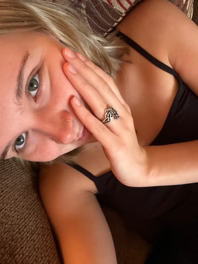 A woman reclines on patterned fabric, possibly a cushion or couch. She wears a black tank top and has fair skin and blonde hair. Her right hand, adorned with a silver ring in the shape of a lion, partially obscures her face. Her eyes are wide and green. A small silver hoop adorns her left nostril. The image is taken from a low angle, creating an intimate and somewhat disorienting perspective.