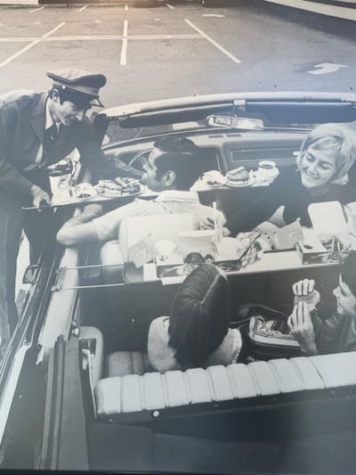 A black and white image shows four people in a car. One person, wearing a cap and what appears to be a uniform, is standing outside the car near the open driver's side door, holding a camera. Inside the car, a woman with short hair is looking out of the window. Two other individuals are seated, with a table between them. The table is covered with various items, including food and drinks. The interior of the car also features light-colored, horizontal-ribbed seats. The scene is located on a road, as evidenced by the white painted lines on the pavement visible on the left side of the image. The specific location within the Township of Langley, Canada cannot be determined from the image alone.