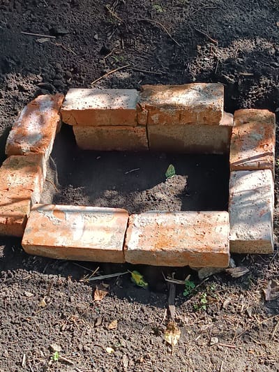 A rectangular structure made of bricks is built on dark soil in an outdoor setting. The bricks are laid in a single layer, forming a border around a central area filled with soil. Some of the bricks appear weathered and have rough surfaces. The soil is loose and contains some dry leaves and small plant debris. The scene is illuminated by sunlight, casting shadows on the bricks and ground. The specific location of this scene in Yasnohirka, Ukraine is not discernible from the image itself.
