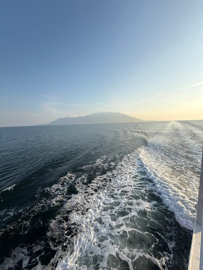 The image displays a wide maritime vista captured from a moving vessel in the waters near Kato Karioutes, Greece. The sky is predominantly clear blue, transitioning to a pale yellow-orange hue along the right horizon. A prominent, dark mountainous landmass stretches across the distant horizon, visible from the left side of the frame towards the center-right. The deep blue-green water in the foreground and mid-ground is marked by a substantial white, frothy wake extending diagonally from the lower right corner towards the center-right, indicating the propulsion of the unseen vessel. A white vertical structure, identified as a component of the vessel, is visible in the extreme lower right corner. No individuals or additional man-made objects are present within the scene.