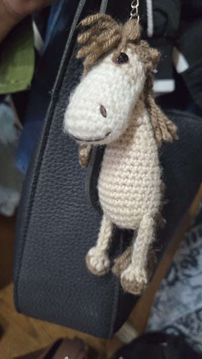 A crocheted plush toy horse lies horizontally on a dark, textured surface, possibly a bag. The horse is primarily beige and white with brown yarn used for its mane and tail. It has simple black button eyes and two small brown stitches for nostrils. The horse is lying on its side, with its legs extended. A silver keychain ring is attached to its head. The background is dark and out of focus, with hints of other objects and fabric. The lighting suggests an indoor setting, possibly during the daytime. There is no visible text in the image.