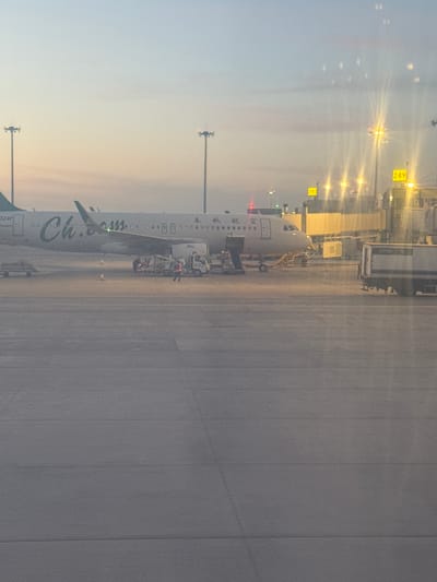 An airplane at an airport, located in Ürümqi, China. The plane is connected to a gate, presumably for passenger boarding or deboarding. The word "China" is displayed on the side of the airplane. Ground crew personnel and vehicles are visible near the plane. The environment suggests dusk or dawn. Lighting fixtures are on.