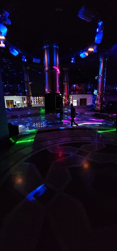 This media file captures a dimly lit indoor scene, likely a club or entertainment venue, with a prominent use of colorful, dynamic lighting. The image is oriented vertically, presenting a somewhat distorted perspective.

The primary subjects are the vibrant light effects, including streaks of blue, pink, and green illuminating the space. These lights reflect off various surfaces, creating a dazzling and energetic atmosphere. Among the objects visible are what appear to be cylindrical structures, possibly part of the venue's architecture or decor, which are brightly lit. In the center of the frame, a silhouetted figure of a person can be seen, their form illuminated by the ambient lights. There are also glimpses of what could be seating arrangements or displays of items in the lower part of the image, featuring multiple rows of what seem to be identical objects.

The overall impression is that of a lively event or a night out, possibly a dance or party, given the energetic lighting and the presence of people. The darkness of the environment suggests it is nighttime. There are no clear geographical or weather cues, but the location context is given as Türkmenbaşy, Turkmenistan. No visible text is discernible in the image. The reflected light and the reflections themselves suggest the presence of mirrored surfaces, adding to the visual complexity of the scene. The lighting creates a mood of excitement and festivity.