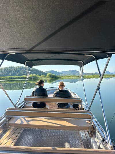 Two individuals are seated in a boat, facing away from the camera, on a calm body of water. The person on the left is a woman with dark hair pulled back, wearing a black leather jacket. The person on the right is a man wearing a straw hat and a dark jacket. The boat has tan upholstery and stainless steel railings. The background features a serene landscape with rolling green hills and mountains reflected in the water. The sky is clear and blue, suggesting a sunny day. This scene is likely captured on Skadar Lake, near Riječani, Montenegro, a region known for its natural beauty and extensive waterways.