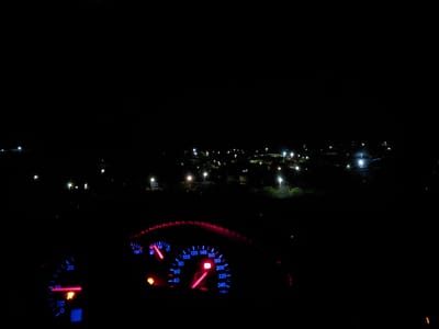 A night scene viewed from inside a vehicle, likely parked on a hill overlooking Dositeevo, Bulgaria. The vehicle's dashboard is visible with lit gauges indicating the car is on, including speed, RPM, fuel, and temperature. Outside the vehicle, there's a nighttime urban view of Dositeevo, with streetlights and scattered residential lights dotting the dark landscape. The horizon is visible as a dark line above the city lights.
