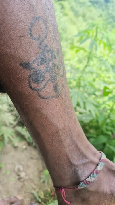 A close-up, horizontal shot shows a person's forearm and wrist, against a blurred background of green foliage and earth. The skin is a dark brown, with visible pores and fine hairs. On the forearm, a black tattoo is partially visible. It appears to be an intricate design, possibly featuring floral or abstract elements. A woven bracelet adorns the wrist. The bracelet is made of thin cords in shades of pink, green, and cream, tied with a pink string. The lighting suggests it is daytime, possibly outdoors. The overall scene conveys a sense of casualness and connection to nature. The location is Shila, India. There is no visible text in the image.