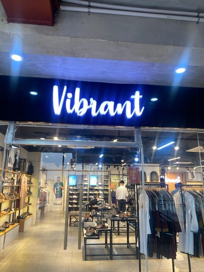 This image depicts the brightly lit storefront of a retail establishment named "Vibrant" located in Narayanganj, Bangladesh. A prominent blue neon sign spelling "Vibrant" is positioned above a clear glass facade that serves as the store's entrance, featuring a central glass door. The overhead exterior structure is exposed, unpainted concrete with visible conduits and recessed white light fixtures.

Inside the store, merchandise is systematically displayed. The left section showcases multiple tiers of wooden and metal shelving presenting various shoe types, including boots and dress shoes, alongside some hanging apparel. A large vertical poster depicting a man in a green shirt is visible further inside this area. The central retail space features several black metal display tables, each exhibiting numerous pairs of shoes, including sandals and slip-ons. The right side of the store, near the entrance, holds clothing racks laden with garments, some individually protected by plastic covers. Behind these racks, additional shoes are arranged on wall-mounted shelves, and several black backpacks are visible on display. A red and white rectangular sign indicating "50%" is affixed to a clothing rack.

Two individuals are present within the store's interior. One person, wearing a light-colored shirt, stands near the back-center, facing away from the camera, seemingly examining merchandise. A second person, partially obscured by the clothing racks on the right, is visible wearing a plaid shirt. The store's interior is illuminated by numerous recessed spotlights, and the floor is covered with light-colored tiles.