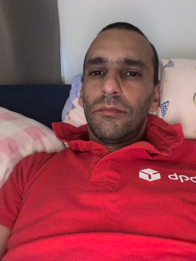 The image is a close-up, vertically oriented, selfie-style photograph of a man lying down. He is wearing a bright red collared shirt with a white logo and text on the collar area. The logo appears to be a stylized cube, and the text above it reads "dop". The man has short dark hair and stubble on his face. His eyes are open, and he is looking towards the camera with a neutral expression. He is lying on a pillow with a blue and white patterned fabric. Behind him, another pillow with a pink and white checkered pattern is visible. The setting appears to be indoors, possibly a bedroom, given the presence of pillows and bedding. The lighting is not strong, suggesting it might be evening or a dimly lit room. No specific location cues are visible to confirm the Schaerbeek, Belgium context, beyond what is provided. The overall mood is relaxed and informal.