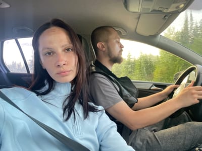 This image captures two individuals inside a car in Ореховая Гора, Russia. A female passenger is seated in the front on the right side of the frame, wearing a light blue, high-neck, quarter-zip top and a dark seatbelt. She has dark brown hair, brown eyes, visible freckles, a gold-colored septum piercing, and a gold-colored labret piercing, and is looking directly at the camera. To her right, a male driver is depicted in profile, holding the steering wheel with both hands. He has a closely cropped haircut, a beard, and wears a gray t-shirt under a dark vest or jacket and gray pants. His eyes are closed or directed away from the camera. The car's interior features a light-colored headliner, an overhead console, and a sun visor. The windshield and side windows are covered with water droplets, indicating rain or recent precipitation. Through the windows, a dense forest of green trees is visible under bright, possibly overcast or late afternoon/early morning, lighting. The steering wheel has a silver-colored center with an automotive logo.