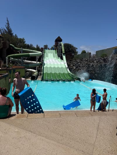 The image shows a scene at the Siam Park water park in Tías, Lanzarote, Spain. A large water slide is prominently featured. A pool of turquoise water is present, and several people are in the water or standing near the edge, holding blue flotation devices. The sky is clear and blue.