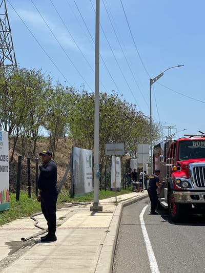 A fire truck is parked on the side of a road in La Playa, Colombia. The fire truck is red and has "BOMBEROS" written on it. Two firefighters are standing near the fire truck, and a third firefighter is standing on the sidewalk. There are also some signs and trees in the background. The sky is blue and it appears to be a sunny day. The fire truck is positioned on the right side of the road, angled slightly towards the sidewalk. One firefighter, wearing a dark uniform and a cap, is standing on the sidewalk, appearing to be observing or directing. Another firefighter, also in uniform, is working near the back of the fire truck. A third individual, possibly a firefighter, is visible near the front of the fire truck. The sidewalk has a hose running across it, which appears to be connected to the fire truck. A few people are gathered further down the sidewalk, near some large signs. The environment is a developed area with a paved road, sidewalk, and grassy verge. The background shows trees and a utility pole with overhead wires.