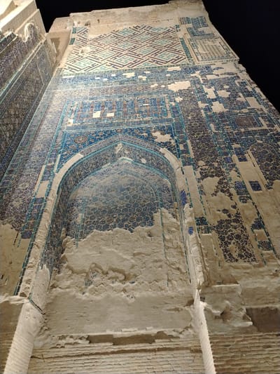 This is a low-angle, ground-level shot of a portion of a historically significant building in Shahrisabz City, Uzbekistan, likely at night given the dark sky. The building's facade is adorned with intricate tilework, predominantly in shades of blue and white. A large arched alcove is visible, also covered in decorative tiles, with some areas of plaster damage and exposure of the underlying brickwork. The tile patterns include geometric designs and possibly calligraphy. The building appears to be constructed of brick, with visible horizontal brick courses at the bottom of the image. The lighting appears to be artificial, highlighting the textures and details of the ancient architecture. No people or other living beings are present in the frame.