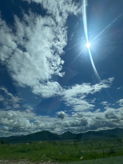 The image displays a wide, landscape view of rolling green hills and mountains under a bright blue sky dotted with white, fluffy clouds. The sun is prominently visible in the upper right quadrant of the image, emitting strong rays and lens flare. The foreground consists of a grassy field, with some patches of dirt and scattered debris. A wire fence is faintly visible separating the field from the distant hills. The scene appears to be taken from a moving vehicle, as evidenced by the reflection on the surface of the lens in the upper portion of the image. The location is identified as Теплоозёрск, Russia.