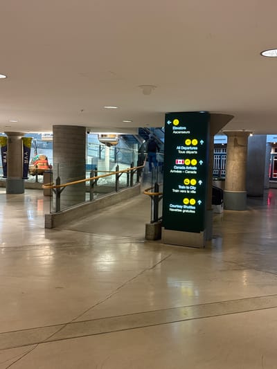 This media file shows a vertical signpost inside a modern building, likely an airport, in Richmond, Canada. The signpost displays directional information in both English and French, with the text "Elevators / Ascenseurs", "All Departures / Tous départs", "Canada Arrivals / Arrivées - Canada", "Train to City / Train vers la ville", and "Courtesy Shuttles / Navettes gratuites". Each directional instruction is accompanied by an arrow and an icon, such as an elevator symbol, a Canadian flag, or a person on a train. The lighting suggests it is daytime, but the interior setting means natural light is not a primary indicator. The floor appears to be tiled and somewhat worn, reflecting the overhead lights. In the background, architectural elements like concrete pillars and glass railings are visible, along with a few people walking and some banners with "YVR" printed on them, which is the IATA code for Vancouver International Airport. The overall atmosphere is functional and transit-oriented.