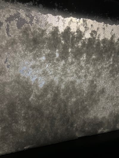 A close-up view shows a surface covered with a grainy, frosted or crystallized substance, possibly ice or snow. The lighting creates highlights and shadows, accentuating the uneven texture. The lower part shows some sort of frame on the right. A dark edge is visible along the frame on the left. The overall color palette is predominantly shades of grey, white, and black, with hints of light blue showing through in the center.