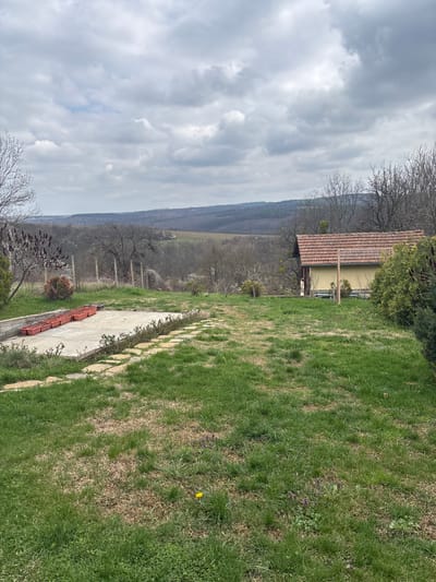 The image shows a landscape in Kavlak, Bulgaria, under a cloudy sky. A yellow house with a red tile roof is visible on a hillside. The foreground features a lawn with patches of brown grass and interspersed with purple flowers and a single yellow dandelion. A stone pathway runs alongside the lawn. The background displays a valley with trees and distant hills.
