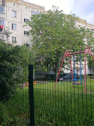 In Khorol, Ukraine, a multi-story apartment building with a rough, concrete facade stands behind a green wire fence. A large tree with dense green foliage is situated in front of the building, partially obscuring the windows. A red and blue swing set is visible in the foreground, positioned on a grassy area. A dark-colored SUV is parked behind the fence near the apartment building. Red clothing hangs from a line outside one of the windows on the left side of the building. The sky above is overcast.