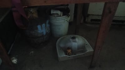 The image shows a cluttered storage area in Putucual, Venezuela. Underneath a wooden table or structure, there is a rusted metal barrel with a faded blue marking, a white plastic bucket containing plumbing parts, and a grimy ceramic sink. The lighting is dim, highlighting the worn and aged condition of the objects. The environment appears unkempt and utilitarian. There are no people visible in the image.