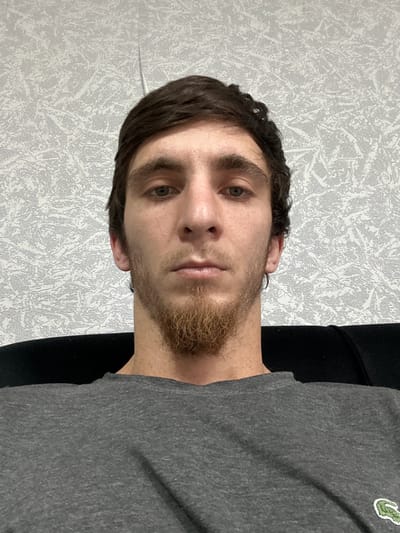 A medium-shot, head-and-shoulders selfie of a young man with dark hair and a light beard. He is wearing a grey t-shirt with a green crocodile logo on the left chest. He is seated in front of a patterned grey wallpaper. His eyes are open and looking at the camera. The image is taken in Chontaul, Russia.