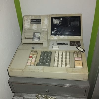 The image shows an old cash register with a black and white display. It has a black key tag attached to the side. The register sits on a drawer unit. It is likely situated in a retail store in Biedenkopf, Germany, based on the provided coordinates. The cash register is dirty and appears to be unused. The cash register has a SHARP label. The image was captured at a low angle. The cash register is white with a number pad and various buttons. The display shows the word "SHARP" and "CASH REGISTER SYSTEM". The cash register is positioned in the center of the image. There are no people in the image. The environment is a retail space, likely a small store.