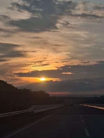 This image captures a sunset over a highway in Seulimeum, Indonesia. The sky is filled with dramatic clouds, illuminated by the setting sun which is partially obscured by darker clouds in the foreground. The sun's rays cast a warm, orange glow across the sky. A silhouetted hillside with trees lines the left side of the highway. The highway itself is dark asphalt, with visible lane markings. A concrete barrier runs along the left side of the road, and another barrier or structure is visible in the distance on the right. A single car is seen driving away from the viewer in the center of the highway. The overall impression is of a transitional moment, with the day giving way to night.