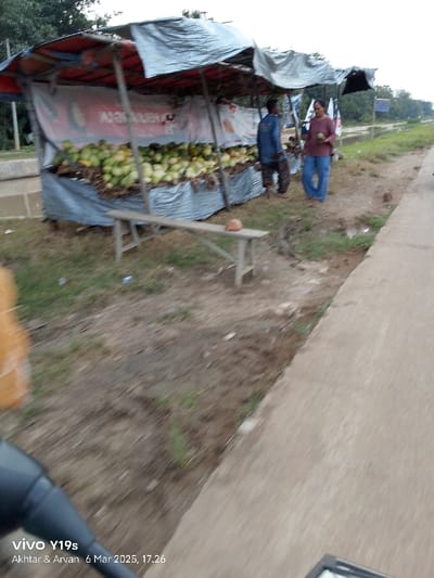 The scene depicts a roadside fruit stand located in the Kecamatan Gabuswetan area of Indonesia. The stand is made of a simple frame with a blue tarp stretched over it to create a shaded shelter. The frame is supporting a large quantity of green fruits, likely melons or watermelons. Two men stand near the fruits, and one woman stands on the opposite side of the stand. There is a wooden bench with a brown object on it, possibly a coconut or a hat. The image appears to be taken from a moving vehicle, as the foreground is blurred and the camera is pointed slightly downward toward the ground.