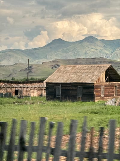 The image shows a rural scene with a weathered wooden barn and other rustic structures in the foreground, set against a backdrop of rolling green hills and mountains under a cloudy sky. A wooden fence is visible in the immediate foreground, partially obscuring the view of the buildings and the grassy field. The barn has a corrugated metal roof and appears to be in disrepair, with visible wooden beams and gaps. Another long, low wooden structure with an open side is situated to the left of the barn. A utility pole stands between the two buildings. The environment is natural and undeveloped, suggesting a remote or agricultural setting. There are no people or animals visible in the image. The lighting is somewhat diffused due to the overcast sky, creating soft shadows.