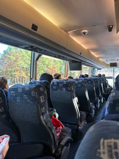 The image depicts the interior of a passenger coach bus, viewed from near the front towards the rear. Approximately 15-20 individuals, primarily adults, are visible, seated in blue upholstered seats with a repeating geometric pattern, arranged in pairs along a central aisle. Several individuals in the middle rows appear to be wearing small, dark head coverings. In the immediate foreground, to the left, a person's hands and legs are visible, with the person holding and interacting with a illuminated smartphone. A red and white striped object is placed on an unoccupied seat towards the middle of the bus.
The bus interior features light-colored overhead storage compartments with integrated strip lighting running beneath them. The ceiling has two dome-style security cameras and one cylindrical camera mounted, along with a small, black rectangular monitor suspended above the aisle. Large windows line the side of the bus, through which an exterior landscape of deciduous trees displaying green, orange, and yellow foliage is visible, indicating travel through a wooded or natural area during autumn. The specific location within Richmond, United States, cannot be determined from the visual information provided.