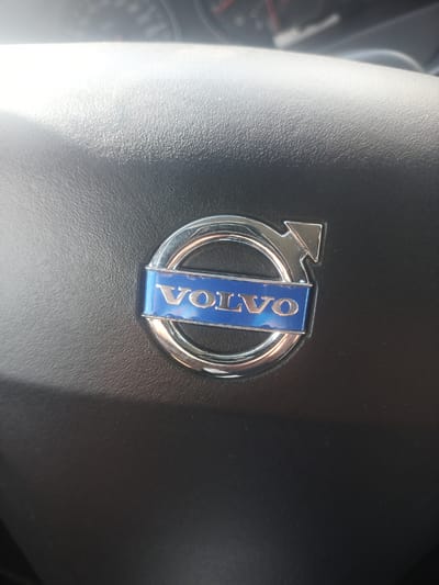 The image is a close-up, eye-level shot of a Volvo logo on a steering wheel, likely in a car. The logo is silver and blue, with the word "VOLVO" clearly visible in silver lettering on a blue background. The steering wheel is dark gray and has a textured surface. Sunlight casts a shadow across the steering wheel, indicating it is daytime. The background is out of focus, but appears to be the interior of a car, possibly including parts of the dashboard. There are no people or specific city details visible in the image.